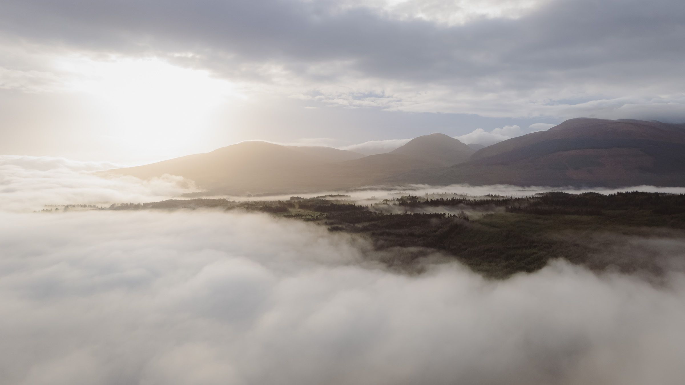 Sun breaking through cloud above a bank of low cloud, with dark forest and layered mountains beyond