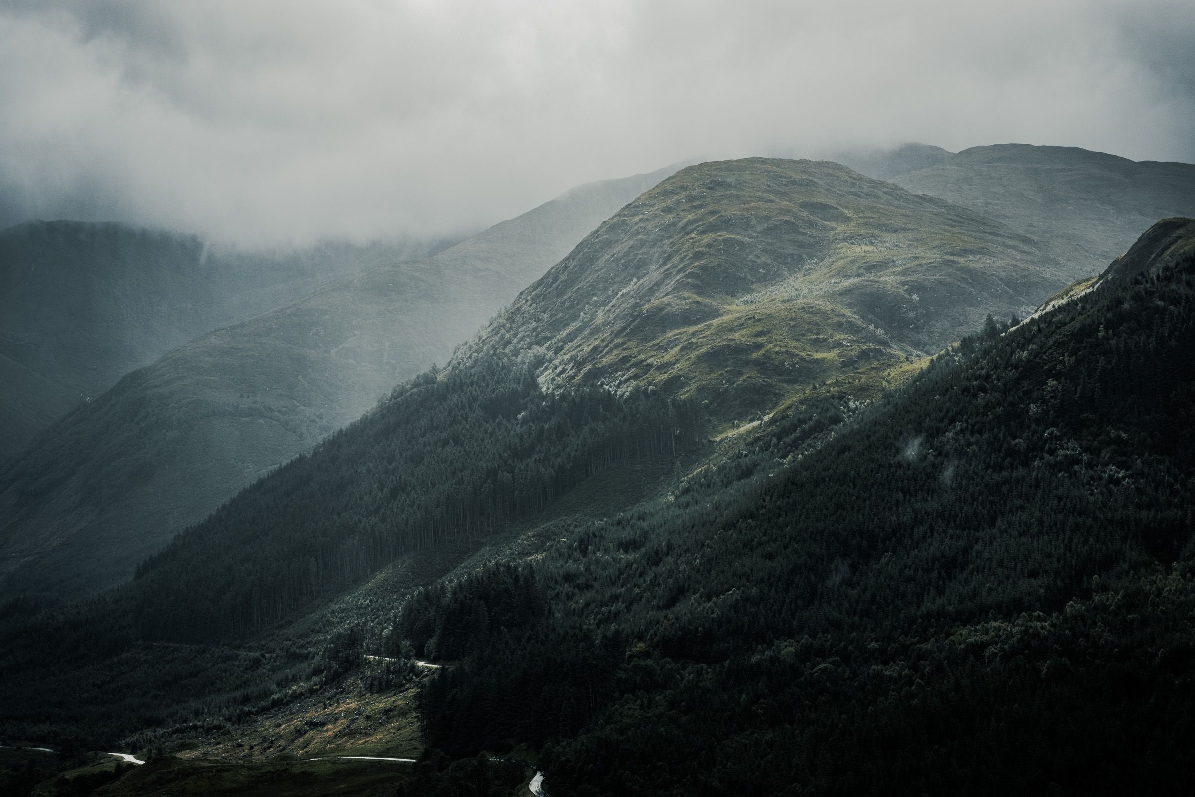 Forested mountain slopes in shifting light under low cloud, with a thin road in the valley below