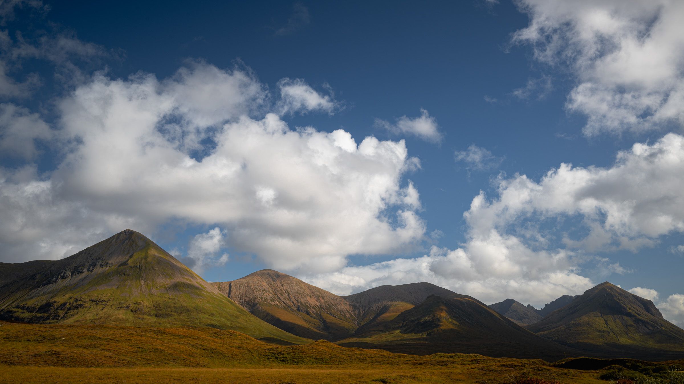 Cone-shaped mountains under a deep blue sky with large white clouds, above golden moorland